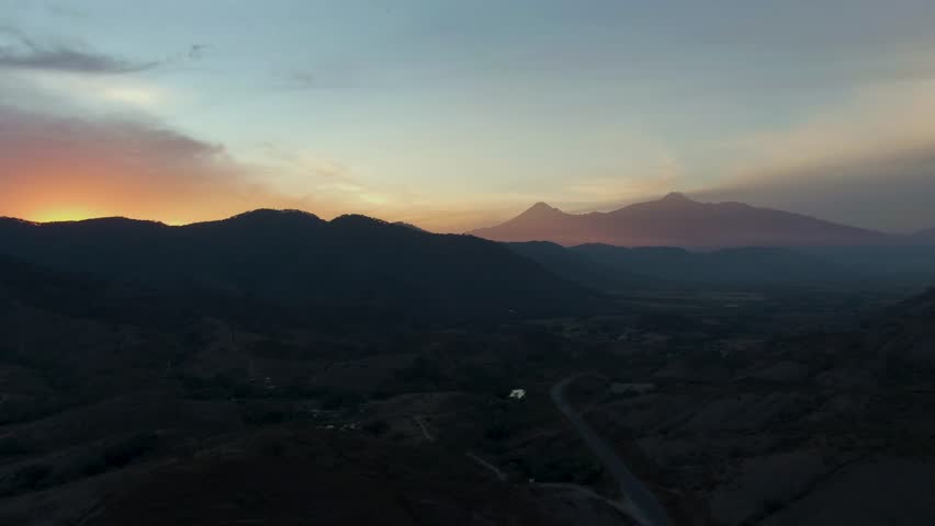 A stunning aerial view captures the distant Nevado de Colima and Colima Volcano during sunset, with dramatic shadows and vibrant hues painting the expansive landscape in rich colors.