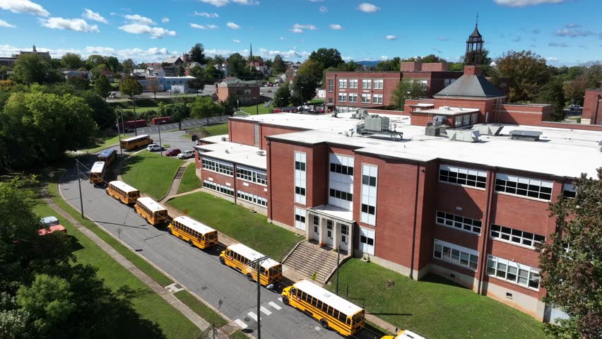 Row of parking yellow school buses along street in front of red brick American school. Aerial backwards wide shot. Sunny summer day in Lynchburg, Virginia.