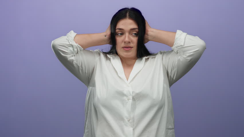 Woman with hands over ears stands against a purple wall, expressing frustration in a white shirt; plus size young lady isolated over plain background, conveying emotion and body positivity.