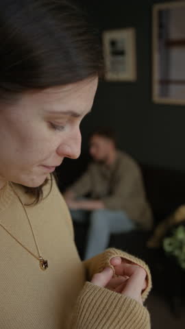 Vertical side view shot of distressed young woman standing by window with young man speaking in blurred background