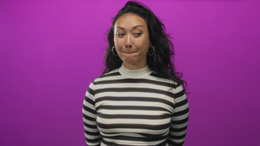 Woman in striped top touching her face with hand and tongue out in purple studio; playful curiosity.
