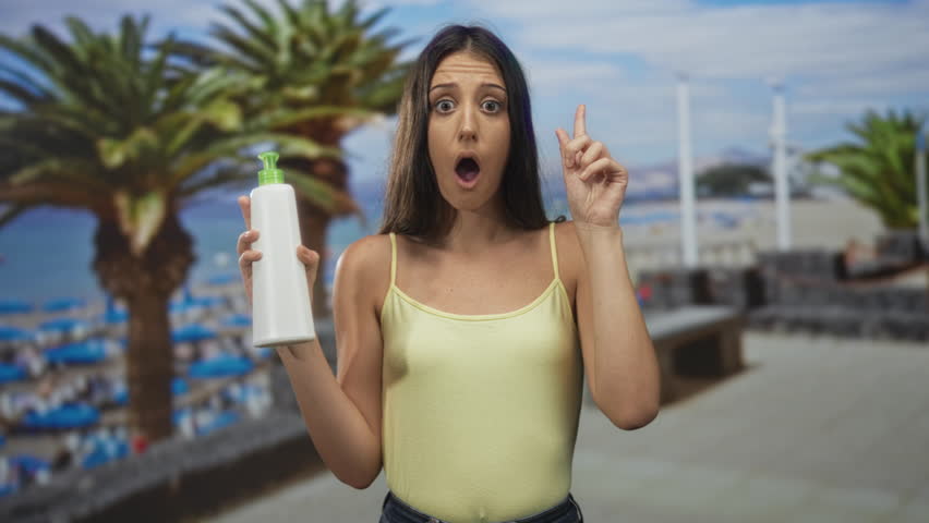 Young hispanic woman holds white pump bottle with one hand and points finger up in studio beach set; product demo surprise.