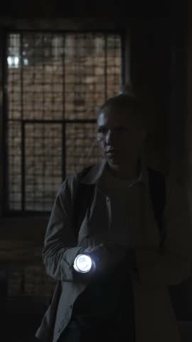 A determined female explorer walks through a dark abandoned corridor, illuminating her way with a flashlight. Mysterious cinematic atmosphere. Vertical video. High quality 4k footage