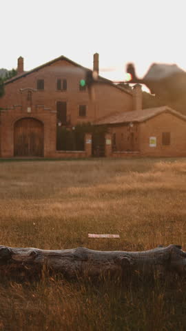 Young man piloting quadcopter drone in a field near a rural house at sunset