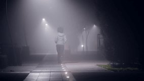 A woman is walking down a sidewalk pavement in the dark foggy time. The street is lit by street lamps. The woman is wearing a white jacket and she is alone. The scene is quiet and peaceful - Powered by Shutterstock - Get 15% off with code: PIKWIZARD15