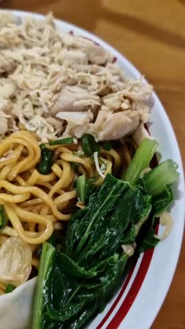 A close-up vertical video of a fork lifting a meatball (bakso) from a bowl of Indonesian chicken noodles, or Mie Ayam, with dumplings.