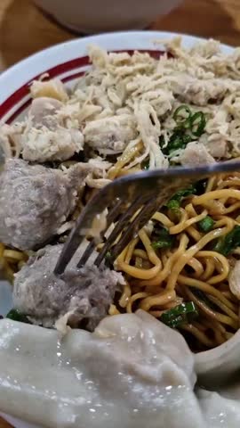 A close-up vertical video of a fork lifting a meatball (bakso) from a bowl of Indonesian chicken noodles, or Mie Ayam, with dumplings.