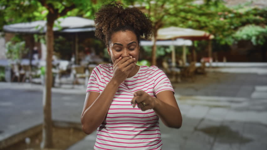 Woman covering mouth with hands laughing at restaurant terrace outdoors in a cafe, striped tshirt and ring visible; surprise unexpected moment.