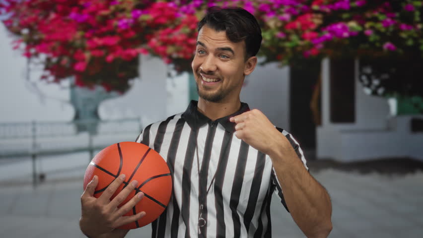 Young hispanic man in referee uniform holding a basketball smiles confidently in a vibrant city street setting surrounded by blooming flowers, capturing the essence of urban sports.