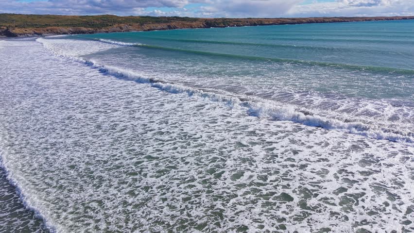 A dramatic aerial sweep across Biscay Bay reveals a curving arc of aquamarine sea meeting white coastal surf under open skies, with sweeping water patterns and distant shoreline.