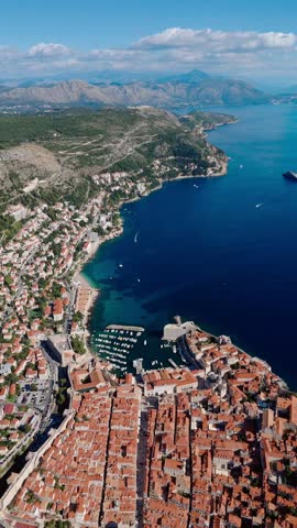 Aerial portrait drone shot flying over Dubrovnik Old Town along the blue Adriatic coast with rugged mountains in the background, capturing the city’s terracotta rooftops and coastal cliffs.