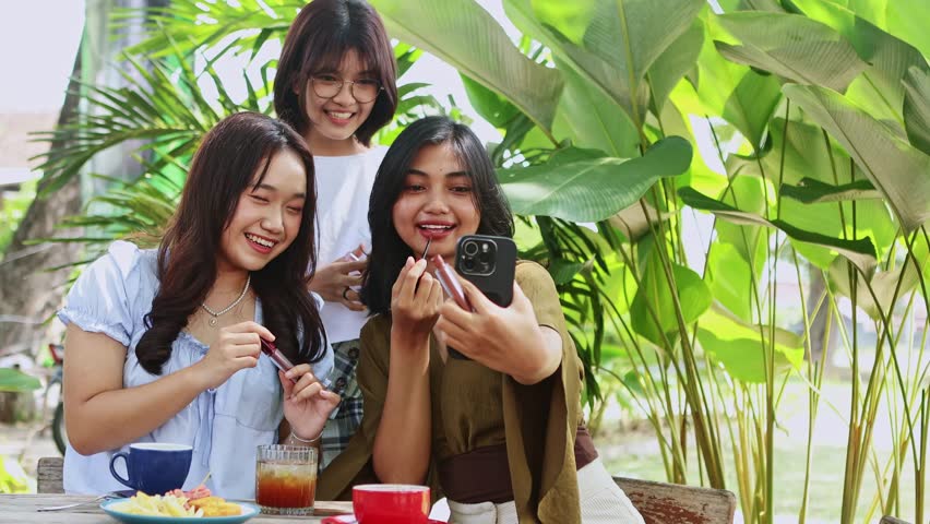 Trendy Indonesian teenage girls bonding at outdoor cafe, applying lipstick and capturing selfie moments with smartphone, showcasing youthful friendship and social media culture
