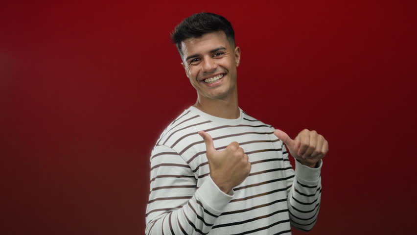 Young man smiling with thumbs up against a red background in a striped sweater, displaying confidence and positivity with an isolated focus on cheerful expression