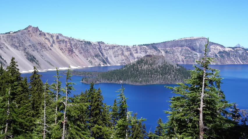 A vivid view of Crater Lake’s deep blue water and Wizard Island framed by tall evergreens under a clear summer sky in Oregon’s Crater Lake National Park.