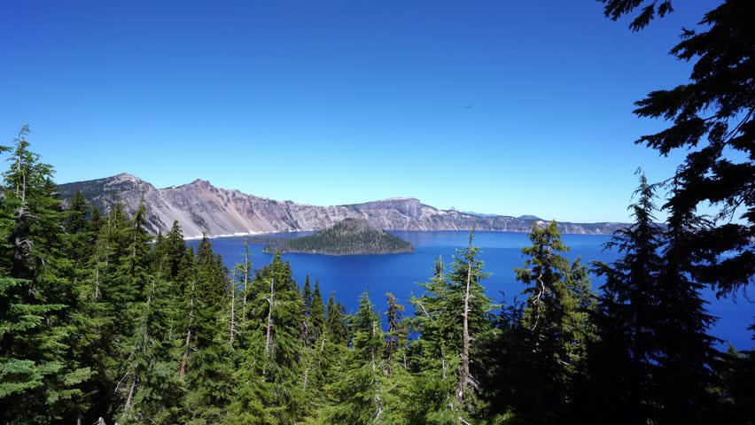 A scenic view of Crater Lake in Oregon framed by pine trees, featuring Wizard Island, blue water, and rugged mountain cliffs under a clear summer sky.