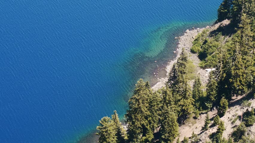 An aerial view shows the deep blue water of Crater Lake meeting the clear rocky shore and pine trees on the steep caldera rim
