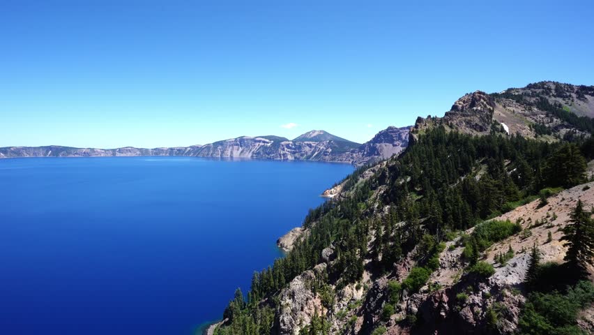 A beautiful landscape shot captures the immense scale of Crater Lake as deep blue water sits below a clear sky and a steep forested caldera rim