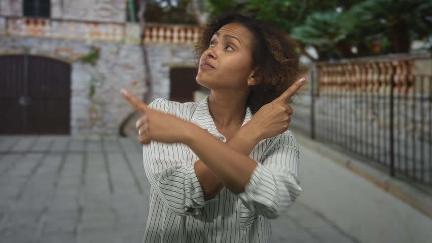 Woman with crossed arms forming an x, index fingers pointing opposite ways on a cobblestone street by a stone building; dismissal uncertainty.