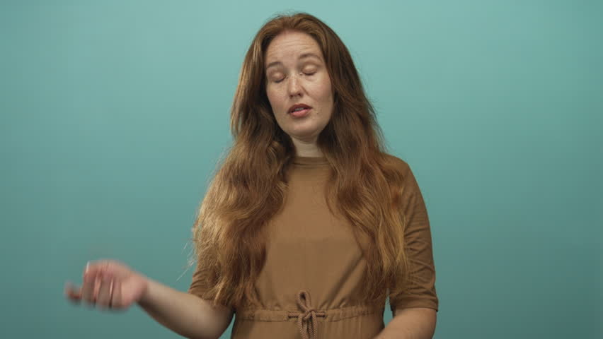 Young redhead woman with hand covers face and crosses arms in studio with teal backdrop; embarrassment.