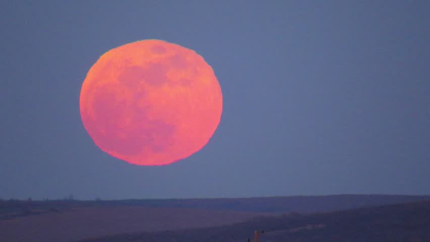 Telephoto shot of a red-orange supermoon rising over a hill at dusk. A white drone with blinking lights passes briefly out of focus in front of the moon. Static cinematic frame. 2021