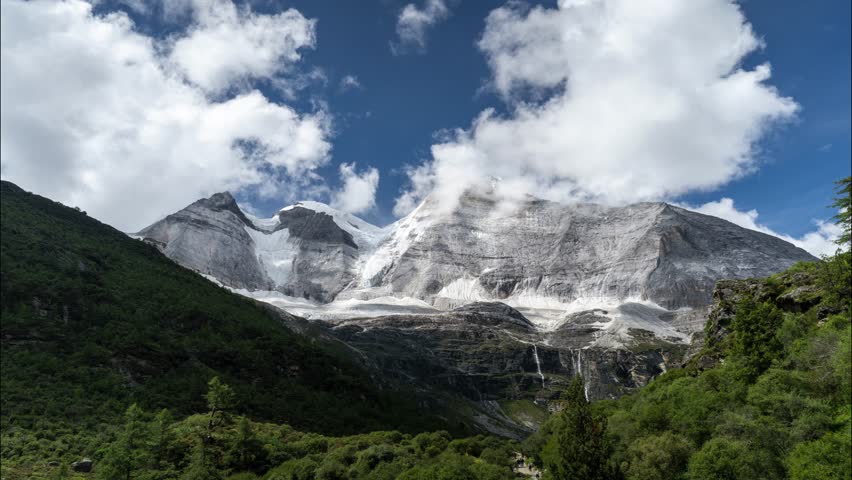 Time lapse view of snow-capped Yading mountains in Inacheng with dramatic clouds moving across blue sky and lush green forest valleys below