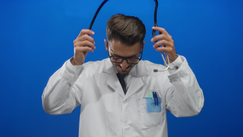 Man doctor wearing glasses smiling adjusting stethoscope in blue studio; professional confidence care wellness.