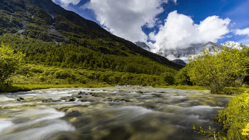 Time lapse view of flowing river through Yading valley with snowy mountain peaks and lush green vegetation in Daocheng, Sichuan Province, China