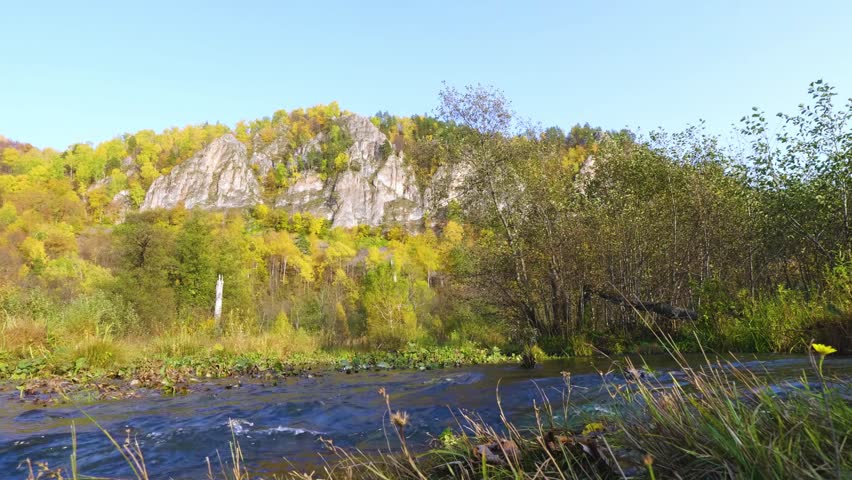 Scenic footage capturing the vibrant autumn foliage on the rocky cliffs and riverbanks of the Sikasya River in the South Ural Mountains of Russia. The clear blue sky and flowing water enhance the natu
