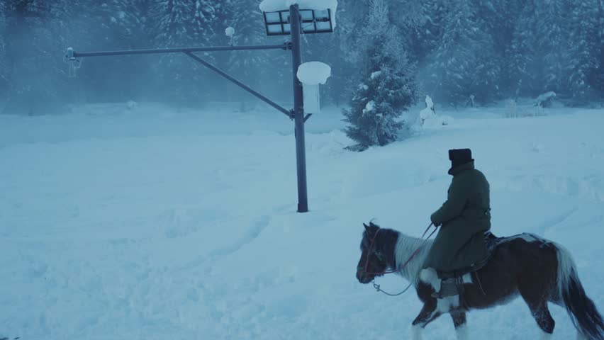 Lone horseback rider traversing snowy winter landscape in misty morning light, creating atmospheric scene with street lamp and snow-covered trees in background.