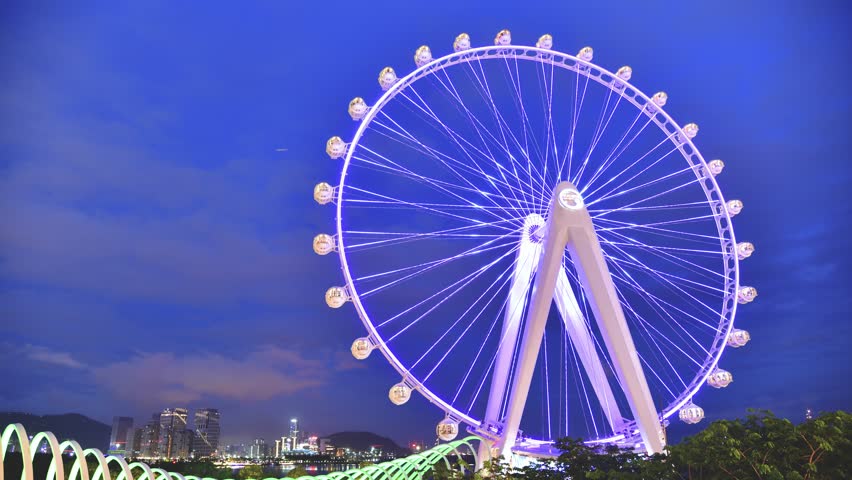 Illuminated ferris wheel against twilight sky with Shenzhen city skyline and bay view, creating stunning urban entertainment landmark scene with colorful lights.