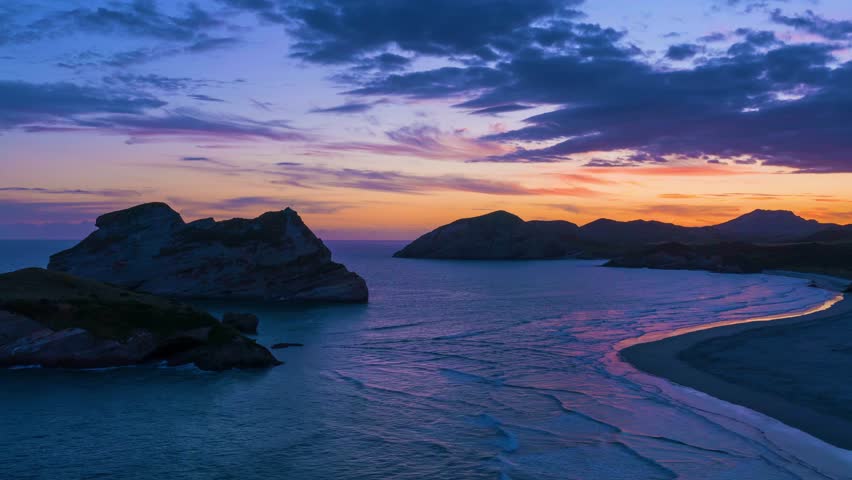 Stunning time lapse seascape with dramatic clouds and colorful sunrise over tranquil ocean channel with rocky coastline and gentle waves washing sandy beach shore