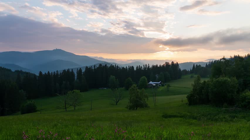 Stunning sunrise time lapse view over green prairie meadows with dense forest and distant mountains creating a serene pastoral landscape with dramatic cloudy sky