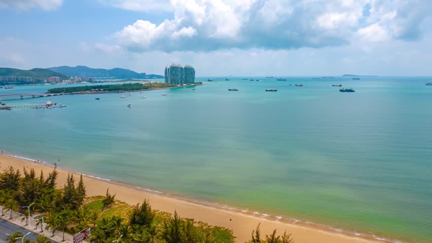 Stunning aerial view of Sanya Bay coastline in Hainan Province, China, showcasing pristine beach, turquoise waters, modern buildings, and dramatic cloudy sky in time-lapse style.