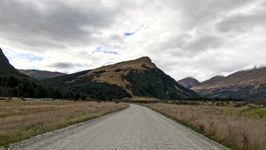 Vehicle travels along rural gravel road with overcast sky, mountains, and grassy fields in view