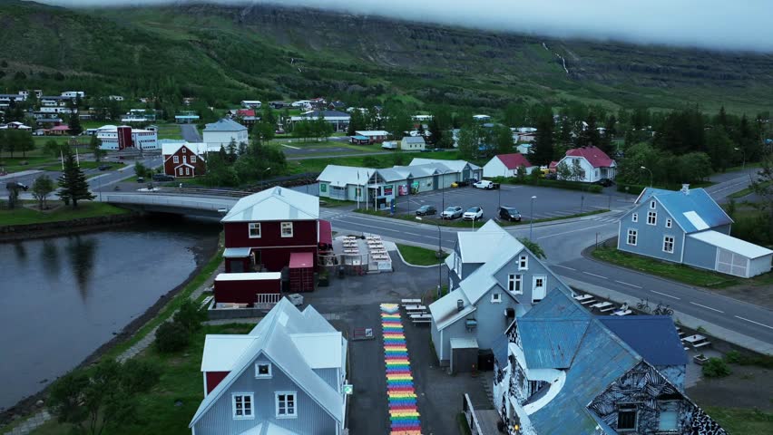 Colorful houses and homes in small Icelandic town during cloudy day. Aerial Birds Eye shot. Seyðisfjörður village with green mountains in distance. Aerial forward wide shot.