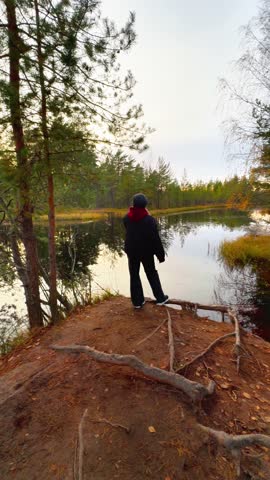 Full shot from a low angle shows a woman wearing a dark jacket and beanie standing on a small, exposed dirt hill overlooking a calm forest lake during a cloudy autumn evening, with exposed tree roots