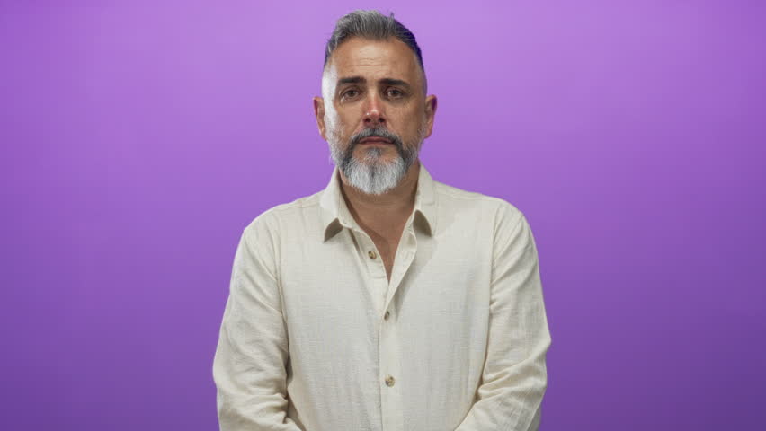 Man, middle aged, with gray beard laughing while seated in a studio with purple backdrop and open collar shirt; joy confidence.