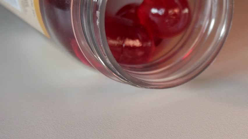 Close-up of red vitamin gummies spilling from a glass jar on a white surface under soft light.