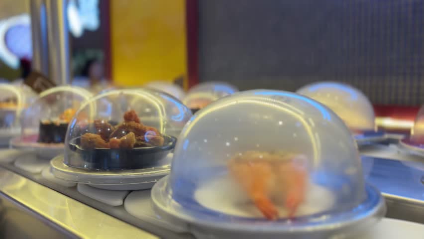 Close up video of sushi plate under a clear dome on a kaiten conveyor in a Japanese restaurant. Soft bokeh, warm light, clean copy space—loopable b-roll for menus, food promos, and social reels