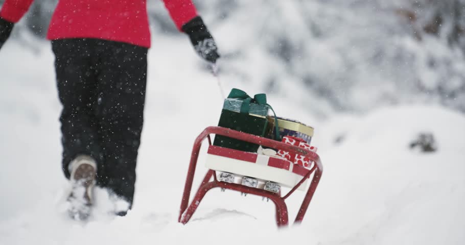 Woman with red winter jacket walks through snow-covered mountain forest while pulling a sledge loaded with Christmas gift boxes