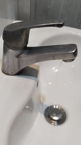 Water drips gently from a shiny stainless steel faucet over a white ceramic sink, shot from the side to show serenity, light reflection, and beauty.