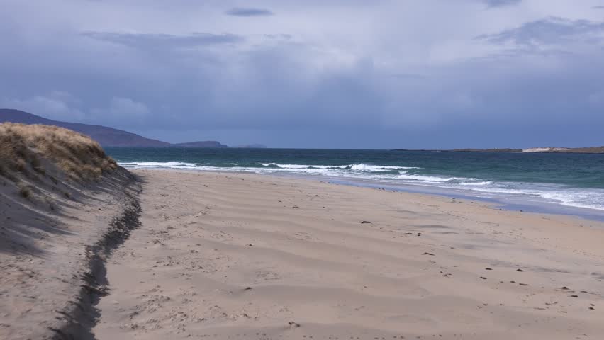Low cinematic drone shot over sun-drenched white sands of an Isle of Harris beach; moody, cloud-wrapped mountains loom dramatically in the background.