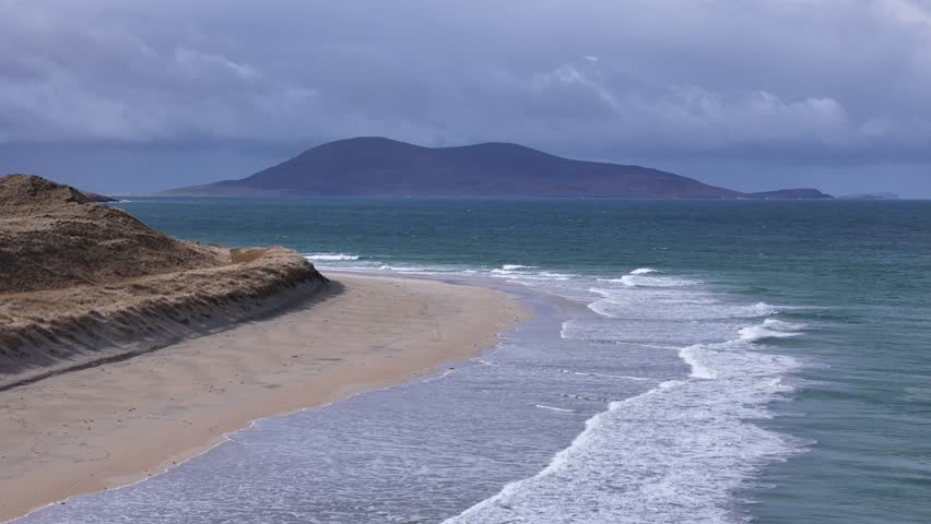 Drone glides backward over Luskentyre’s white sands, revealing moody, cloud-wrapped Harris mountains in a striking sun-and-shadow contrast on Scotland’s wild Atlantic coast.