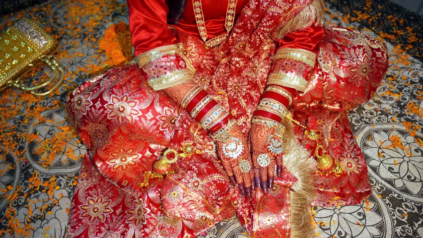 Bride Hands Embellished with Henna and Jewels, Spouse sits gracefully on blossom decorated honeymoon bed, Newlywed woman glows in a crimson golden ensemble, Traditional attire sparkles amid Petals