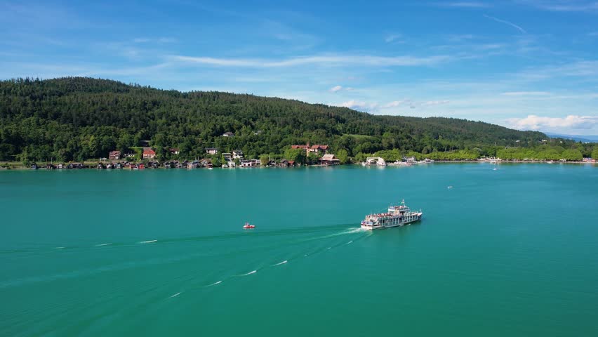Aerial landscape of Worthersee in Klagenfurt, Austria featuring charming waterfront homes, lush greenery, tourist boat and serene blue waters under a clear sky.