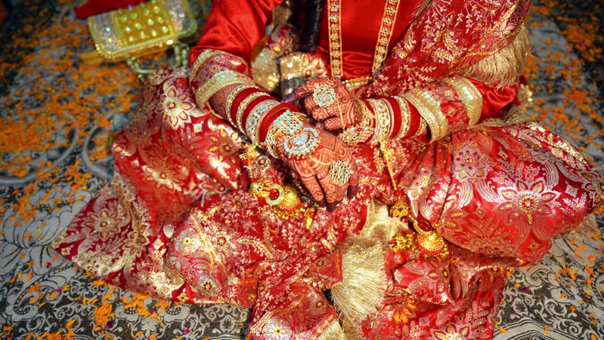 Spouse sits gracefully on blossom decorated honeymoon bed, Bride Hands Embellished with Henna and Jewels, Newlywed woman glows in a crimson golden ensemble, Traditional attire sparkles amid Petals