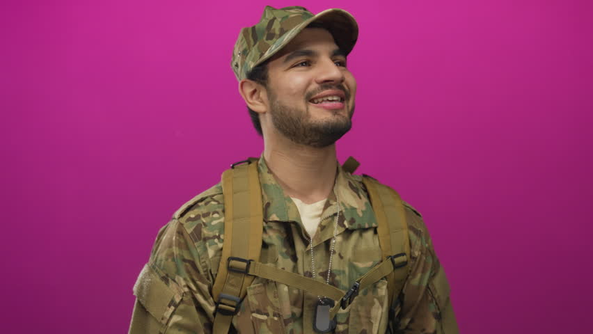 Man in camouflage military uniform adjusts backpack strap and smiles confidently in studio with vibrant purple backdrop; pride.