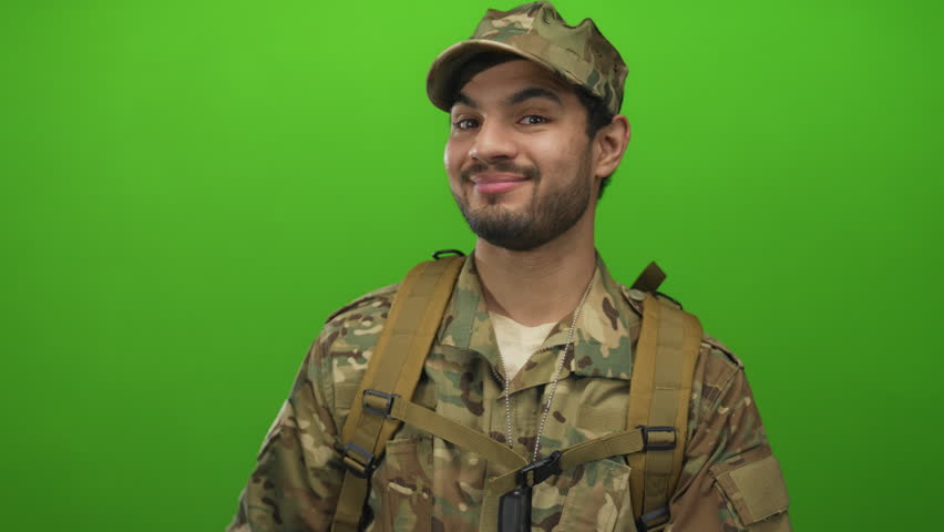 Man in camouflage military uniform wearing tactical backpack points finger with a smile in green studio; pride.
