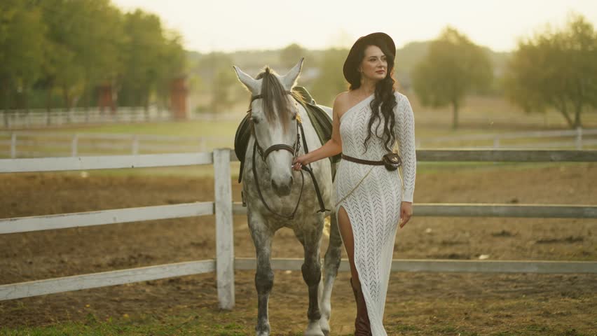 Woman in hat holding reins next to gray horse on ranch. Woman with horse concept.
