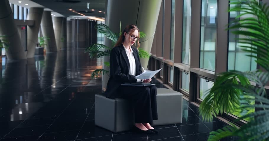 Stylish businesswoman in black suit and glasses sits writing or signing document in brightly lit office hall with sleek modern architecture - slow motion, parallax shot